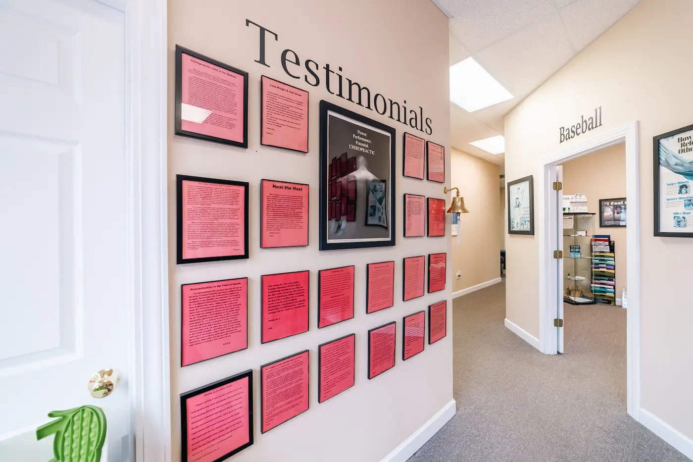 A hallway in a chiropractic office with a 'Testimonials' wall featuring framed documents and a 'Baseball' themed room entrance.