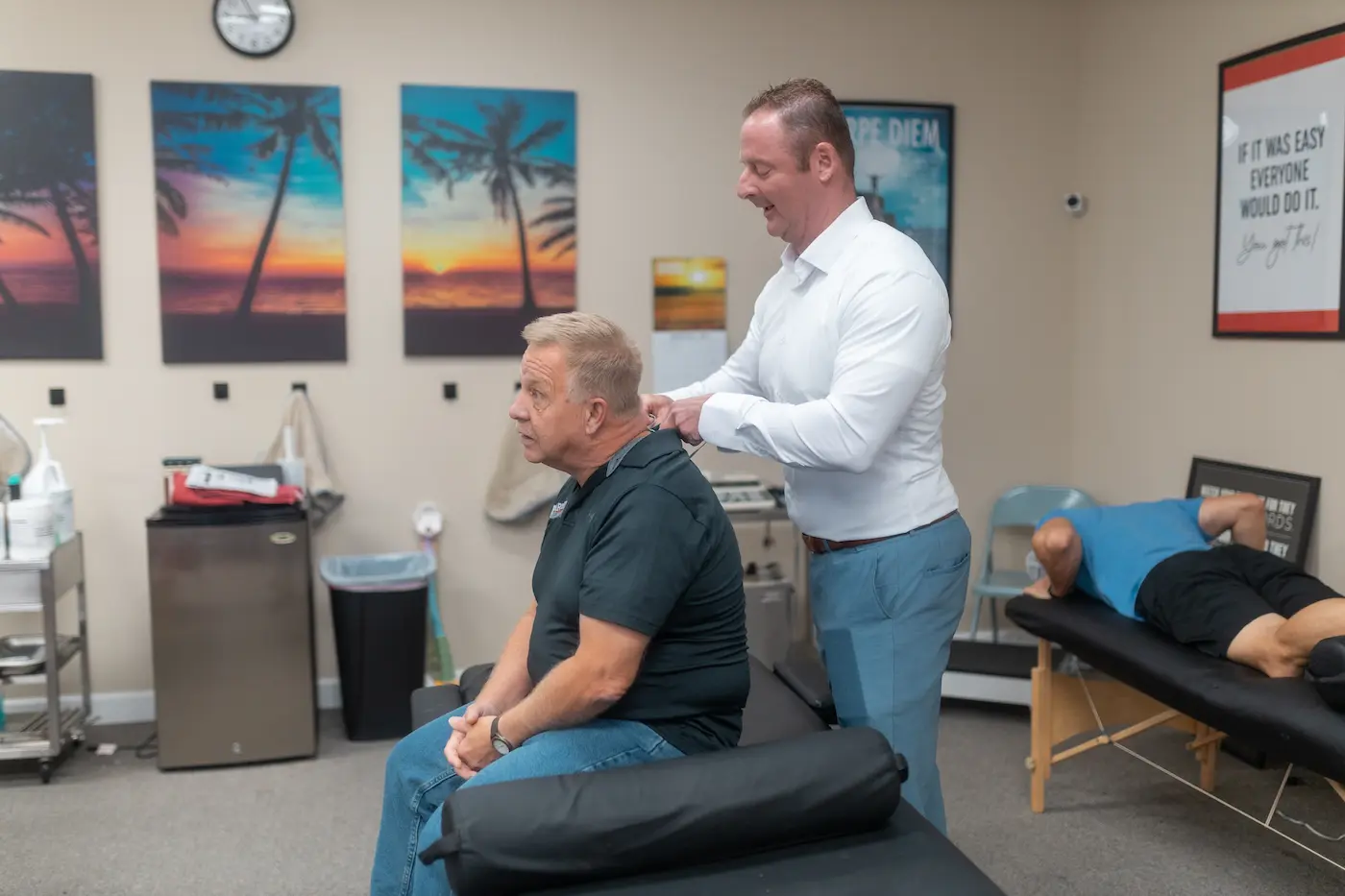 A male practitioner adjusts an older male patient's neck in a clinic. Another patient lies on a table in the background. Motivational posters and palm tree art decorate the walls.