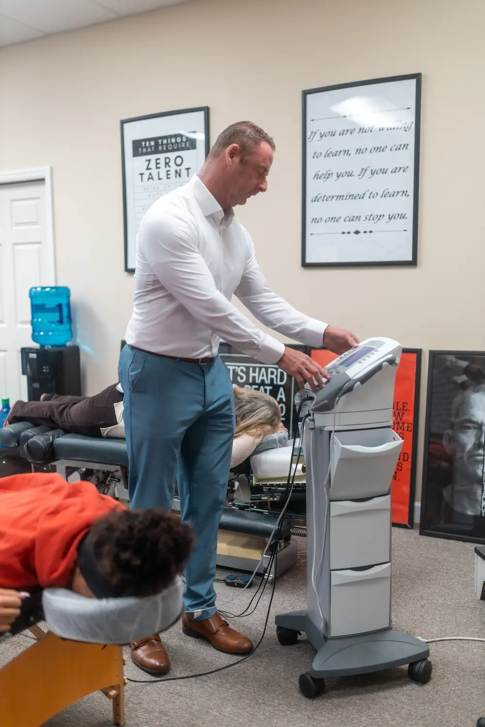 A male practitioner adjusts a medical device while two patients lie face down on treatment tables in a clinic. Motivational posters are on the wall.