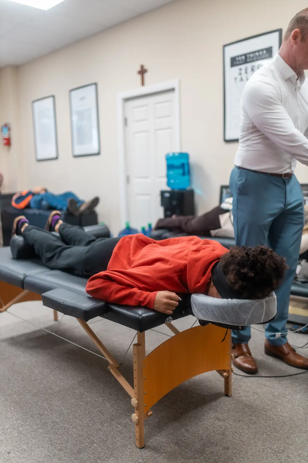 A person in a red hoodie lies face down on a black massage table, with a man standing nearby in the background.