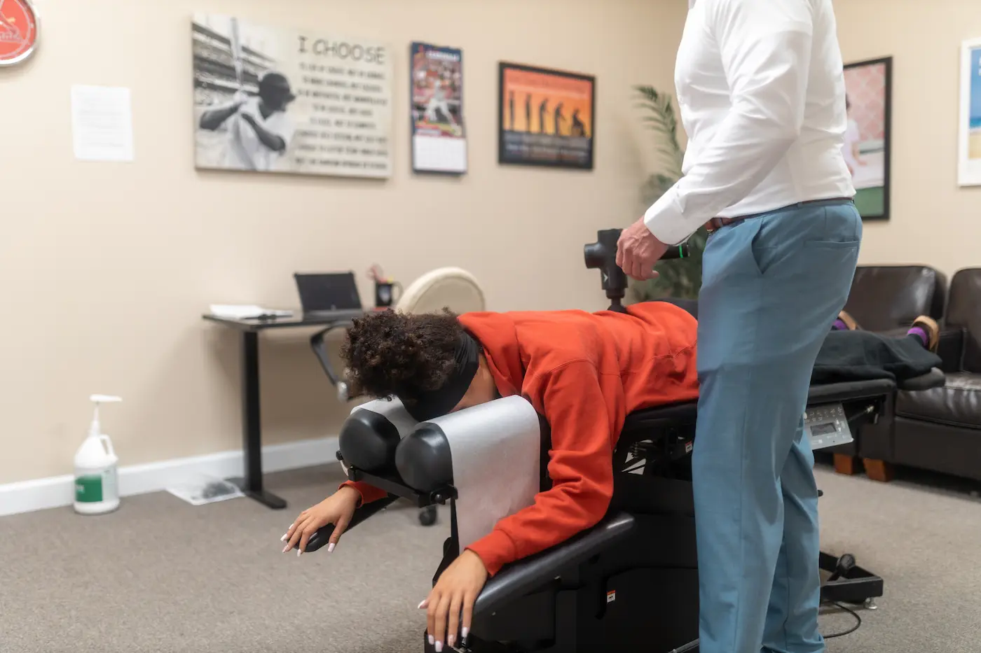 A person lies face down on a chiropractic table while a practitioner uses a massage gun on their back.