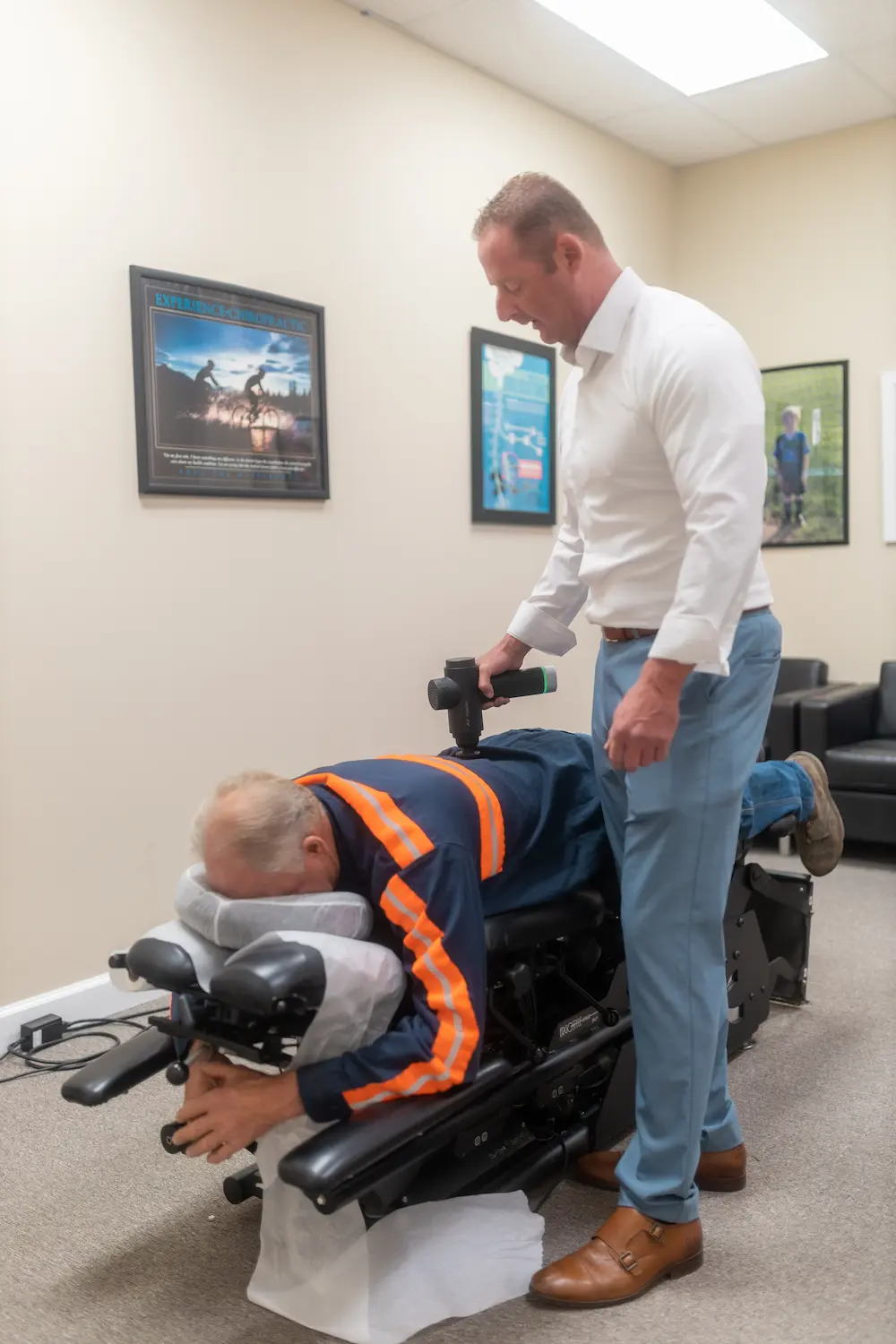 A man lies face down on a chiropractic table while another man applies a percussive massage device to his lower back.