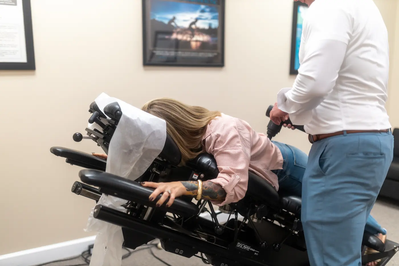 A woman lies face down on a chiropractic table while a man applies a percussive therapy device to her lower back.
