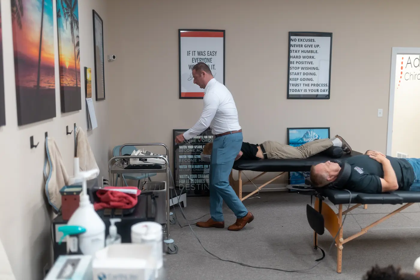 A male practitioner in a white shirt stands near a medical cart in a chiropractic office, with two male patients lying on treatment tables. Motivational posters adorn the walls.