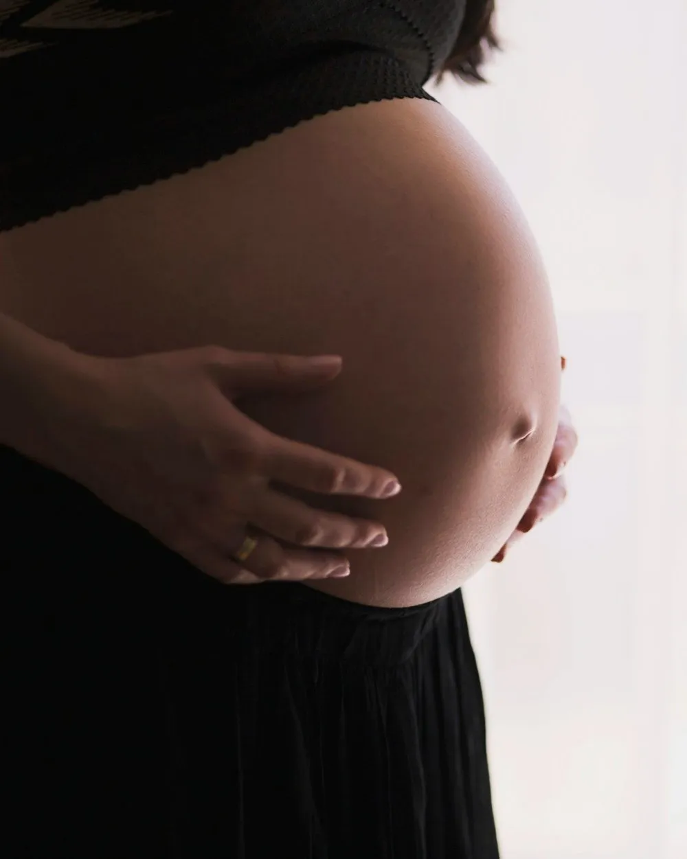 Close-up of a pregnant woman's belly, with her hands gently resting on it. A ring is visible on her finger.