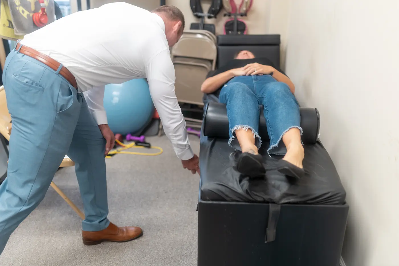 A practitioner in a white shirt and blue pants leans over a patient lying on a black examination table.