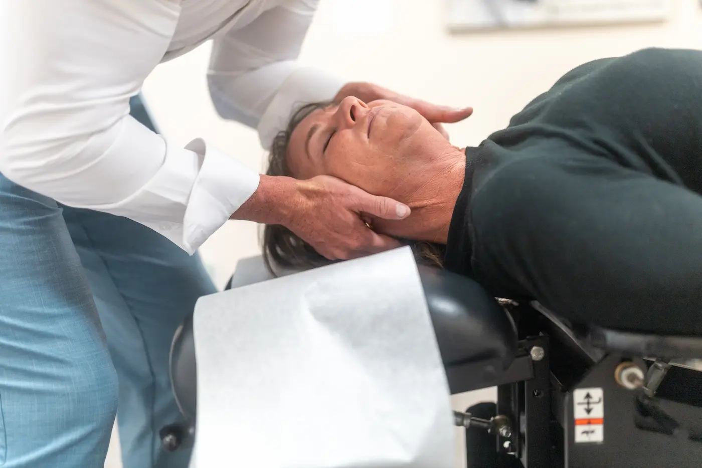 A person lies on a chiropractic table as a chiropractor adjusts their neck with both hands.