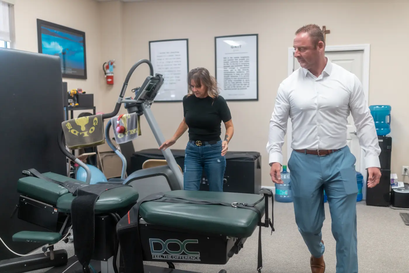 A woman and a man in a physical therapy clinic with exercise machines and dark green treatment tables.