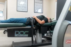A woman lies on her back on a black and green spinal decompression table with a harness around her waist.