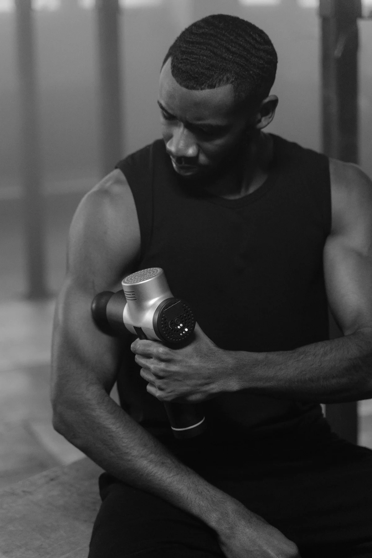 A man in a tank top uses a massage gun on his bicep in a black and white image.