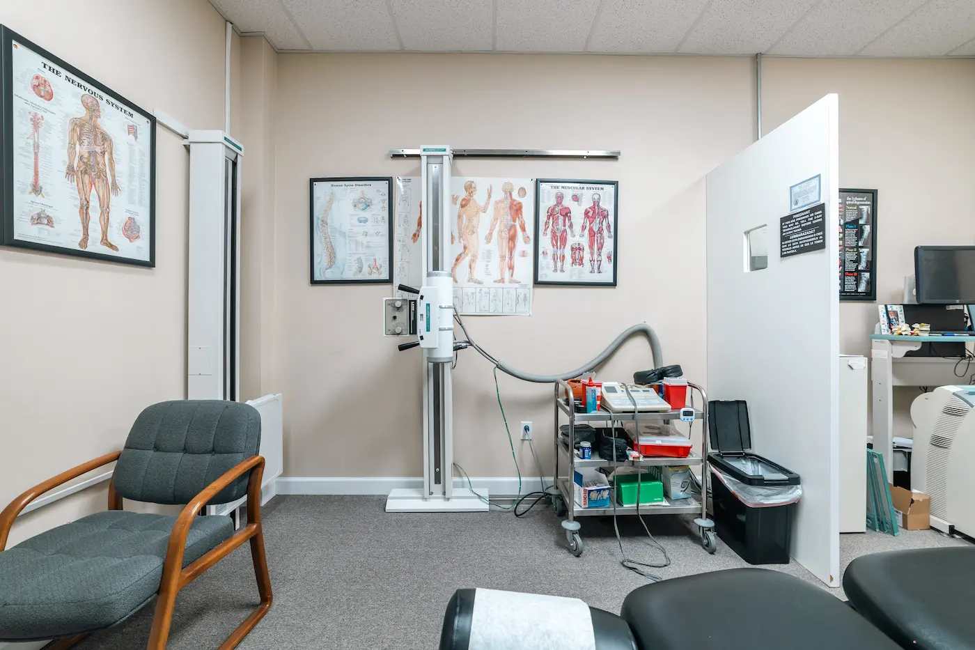 A medical examination room with an X-ray machine, anatomical charts, a utility cart with supplies, and a grey chair.