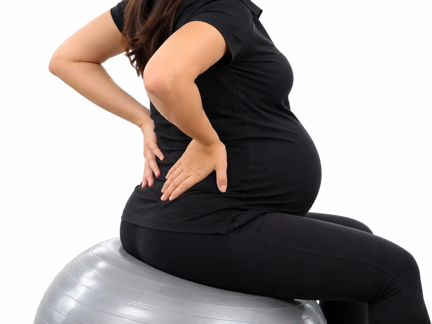 Pregnant woman in black clothing sitting on a gray exercise ball, holding her lower back.