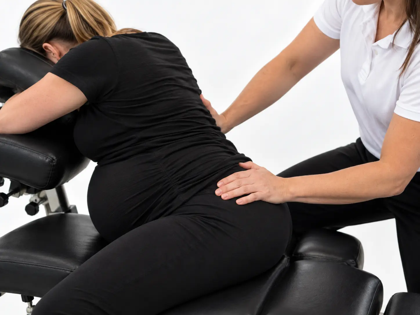 A pregnant woman in black clothing receives a back and hip massage or adjustment from a therapist on a black massage chair.